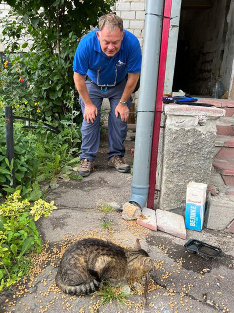 The war in Ukraine: How Naturewatch Foundation has been supporting animals in crisis 5 A smiling Ukrainian man is bending over outside in a garden, watching a cat eating food provided by a feeder in the form of a drainpipe.