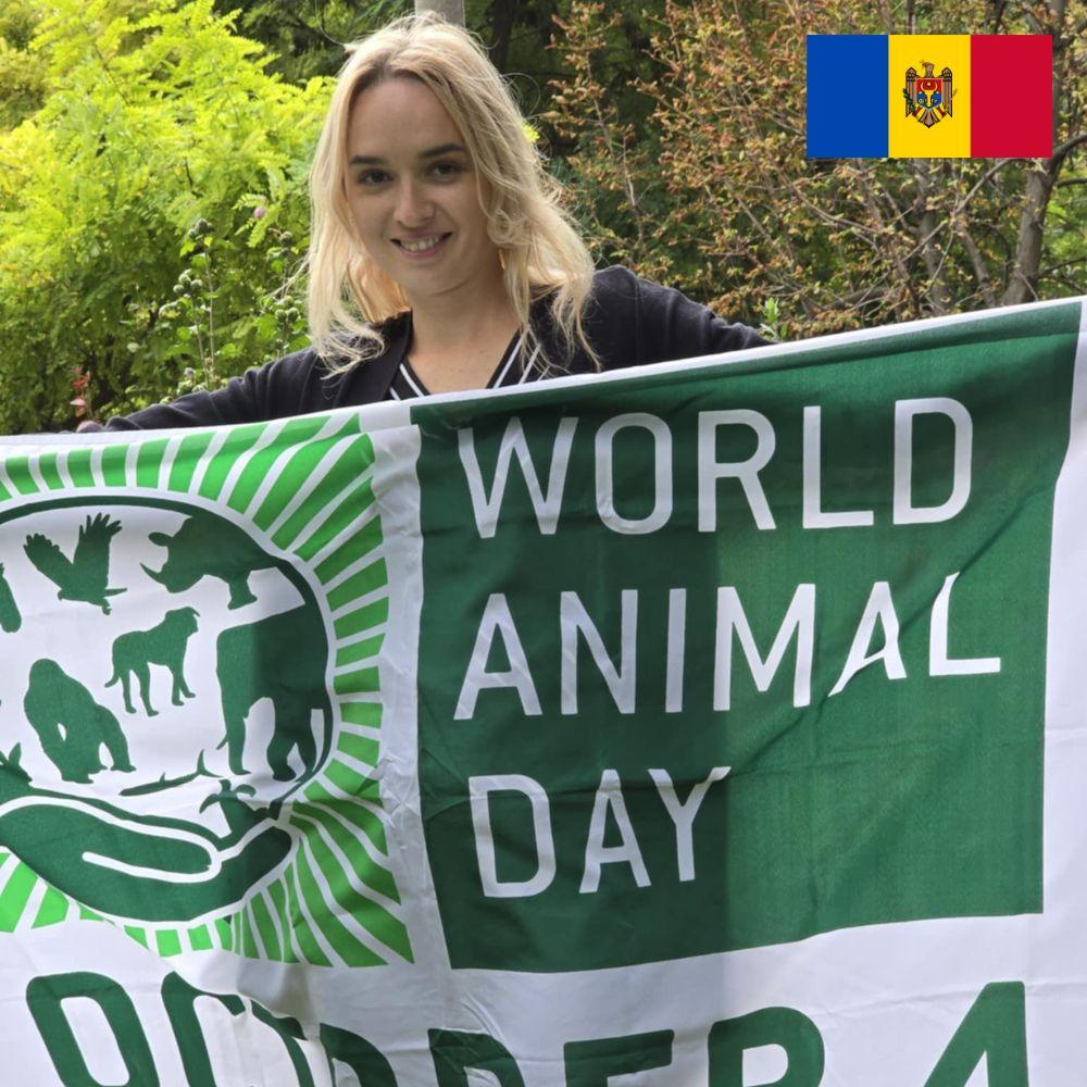 A smiling woman with blonde hair is standing outside, holding a flag featuring a green and white World Animal Day logo. There are trees in the background.