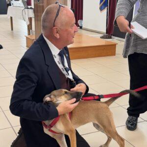Zero to Hero: Police Training Across Greece for Safer Communities and Protected Animals 1 Mark, wearing a suit, kneels on the floor smiling as he pets a tan dog with a red hardness during a training event in Athens, Greece.