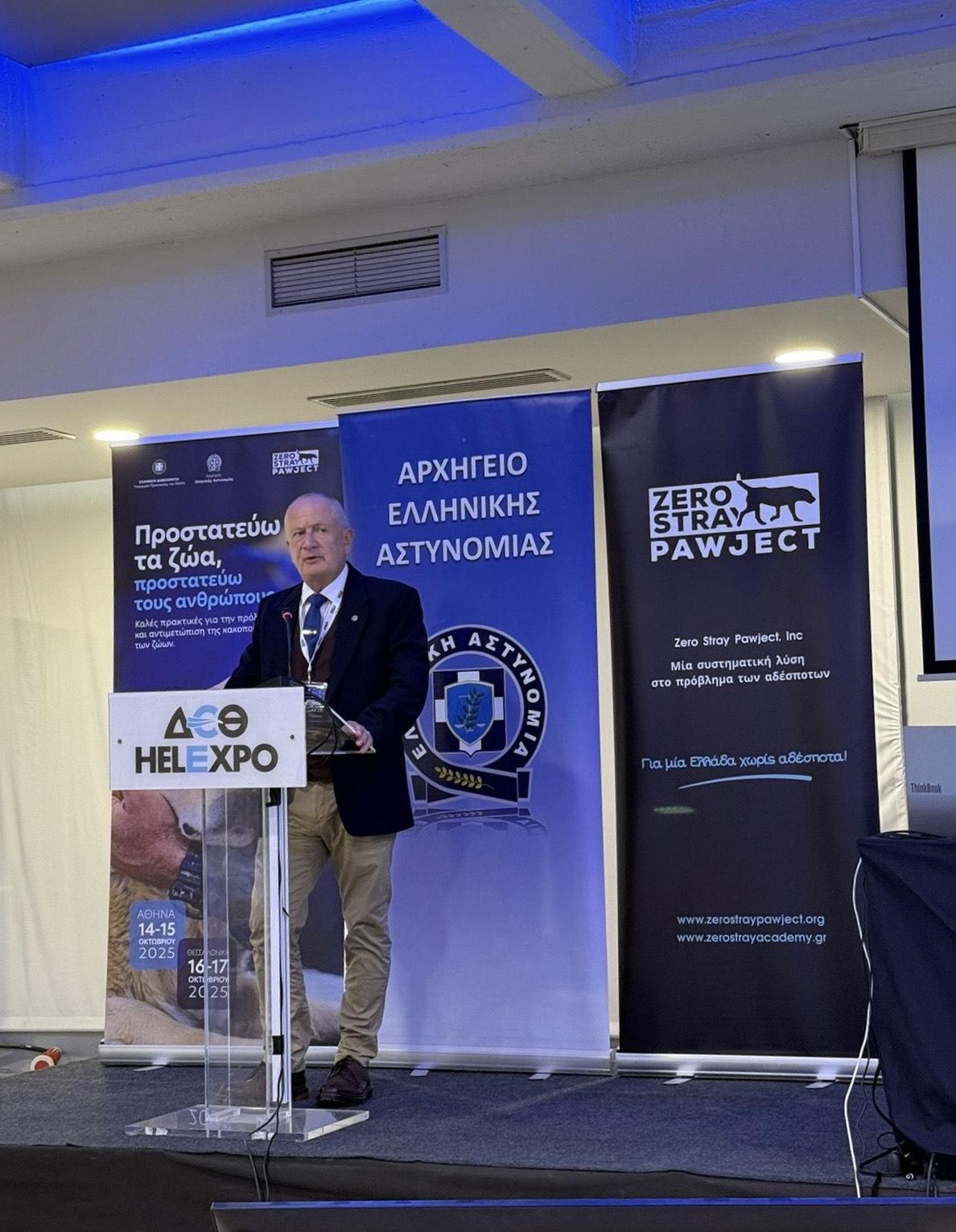 Mark stands at a speakers podium. The background shows a poster about protecting animals and people, as well as the Hellenic Policy and Zero Stray Pawject logos.