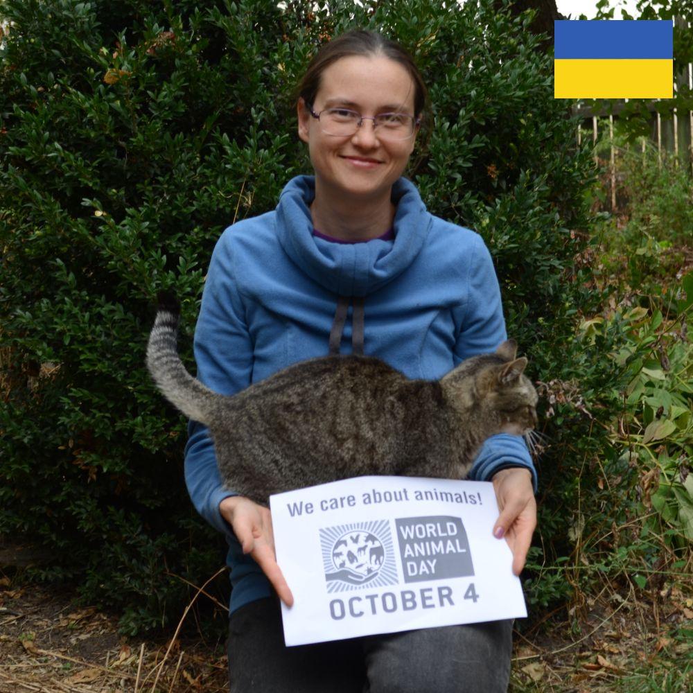 A woman with dark hair is kneeling in a garden, smiling. She has a tabby cat in her lap and is holding a paper sign with a World Animal Day logo and a message that reads, &lsquo;We care about animals!&rsquo;