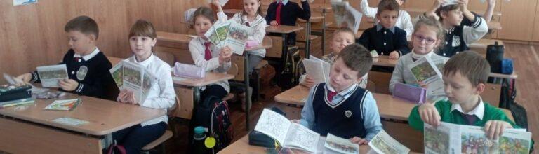 Classroom of primary school students, sitting at desks, holding open picture books that feature animals. The atmosphere is lively and engaged.