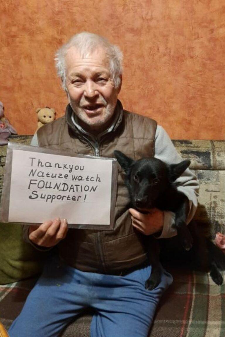 Your support 9 A man smiles while holding a sign saying "Thank you Naturewatch Foundation Supporter!" He’s sitting on a couch, cuddling a black dog.