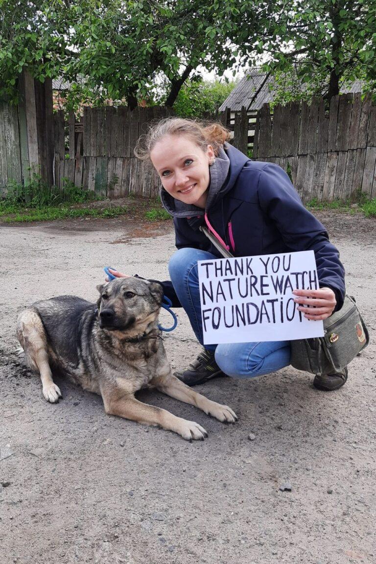 Your support 8 A woman kneels beside a calm, dog who is lying down. She holds a sign that reads "Thank You Naturewatch Foundation." Wooden fence and greenery in the background.