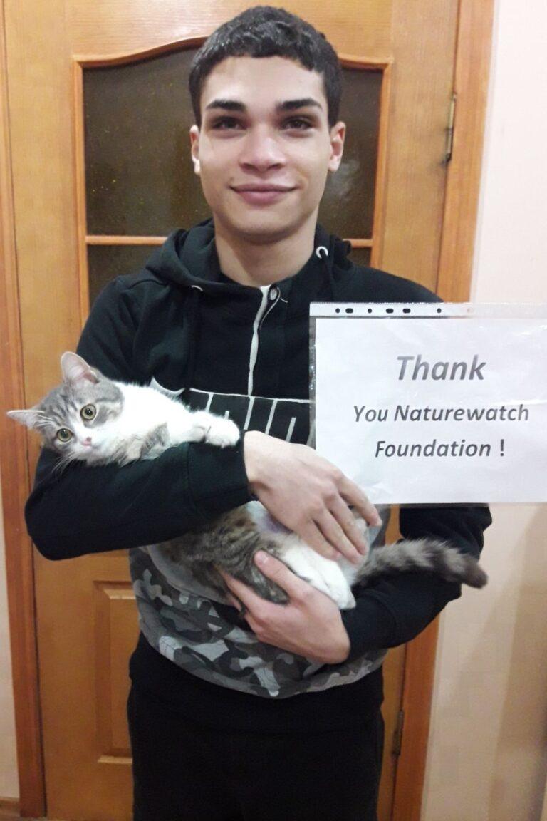 Your support 11 A young man stands in front of a doown door. He smiles while holding a fluffy gray and white cat and displays a paper reading "Thank you Naturewatch Foundation!"