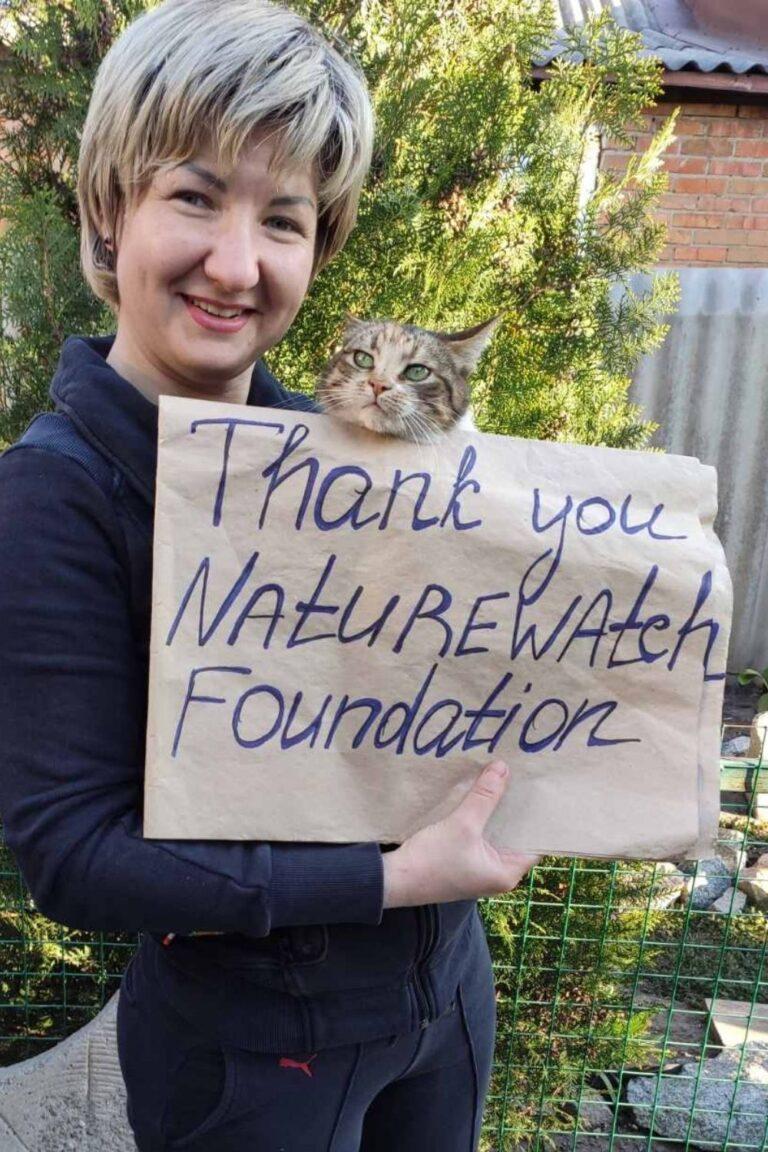 Your support 2 A smiling woman holds a tabby cat alongside a handwritten sign reading "Thank you Naturewatch Foundation." The setting is outdoors, with trees and a brick wall.