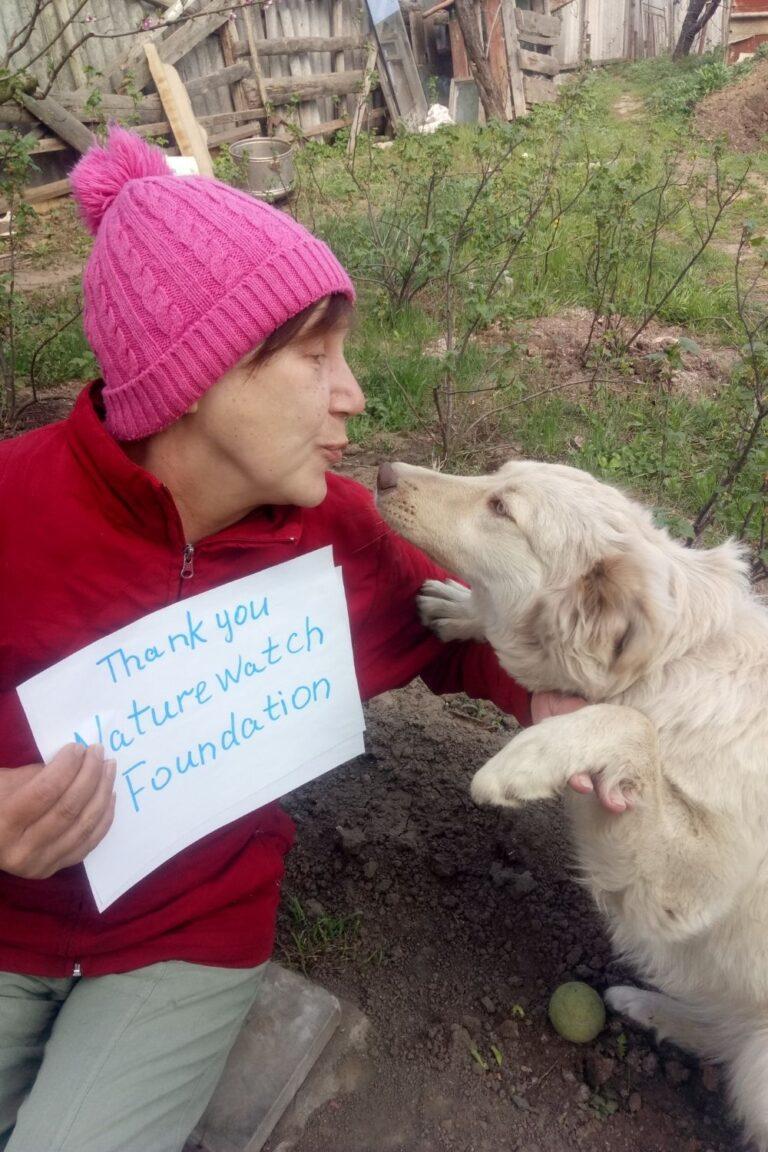 Your support 3 A woman in a red jacket and pink woolly hat shares an affectionate moment with a white dog in a garden. She holds a sign reading "Thank you Naturewatch Foundation."
