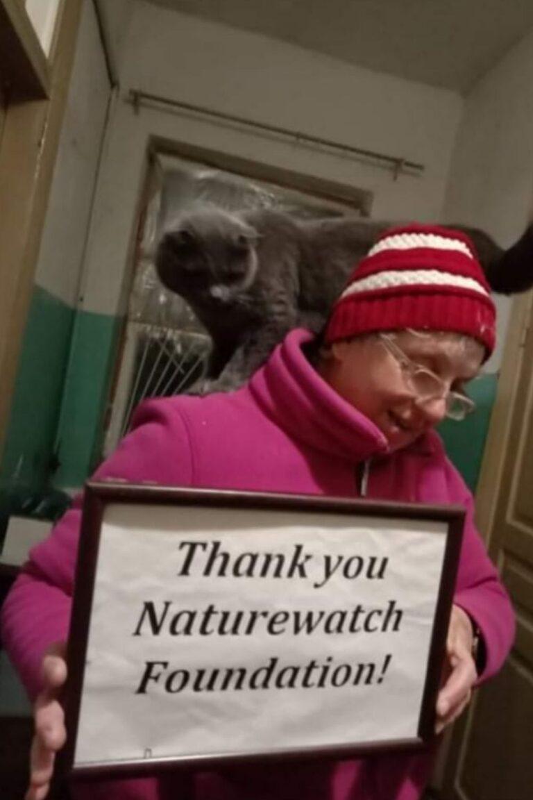 Your support 5 A smiling woman in a red beanie and glasses holds a framed sign reading "Thank you Naturewatch Foundation!" A grey cat is playfully perched on her shoulders.