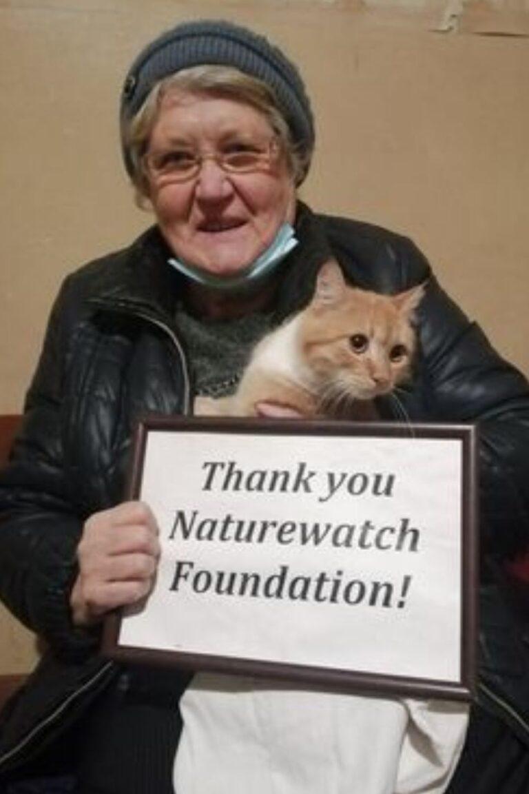 Your support 13 A smiling woman in a blue hat and glasses holds a ginger and white cat and a framed sign reading, "Thank you Naturewatch Foundation!"