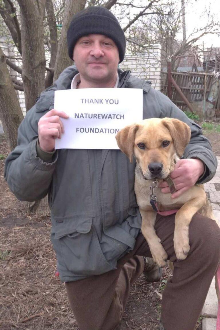 Your support 7 A man in a winter jacket and beanie holds a paper sign reading "Thank You Naturewatch Foundation" while cuddling a brown Labrador-type dog. Outdoor setting with trees.