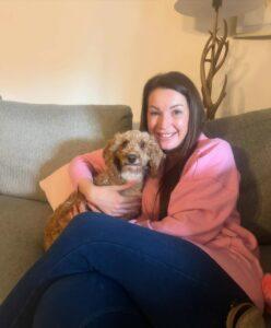A woman in a pink jumper and blue jeans sitting on a sofa with a small brown and white cockapoo dog.