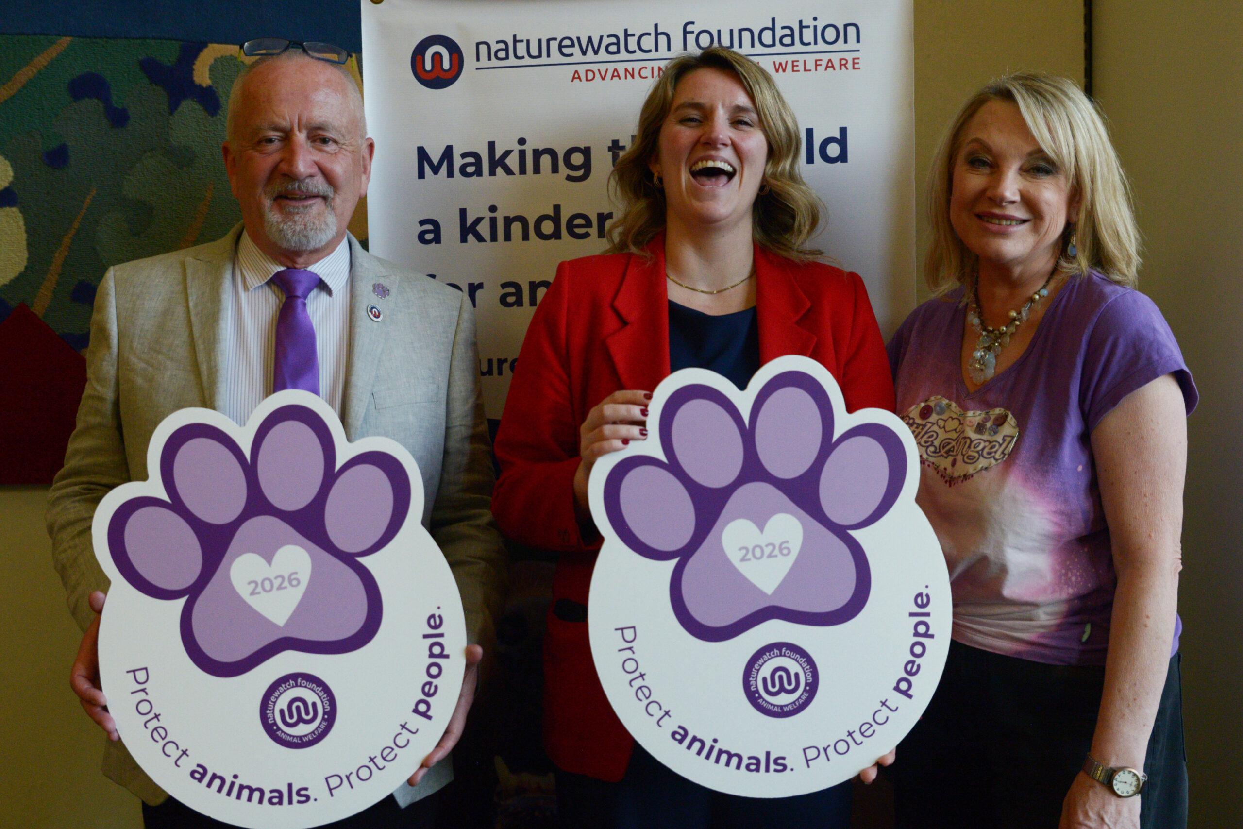 Cross-Party Support for Purple Paw at Parliament 1 From left to right are Mark Randell, Wendy Turner Webster, and MP Jess Brown-Fuller. They are smiling and each holds a large purple paw-print sign reading “Protect animals. Protect people."