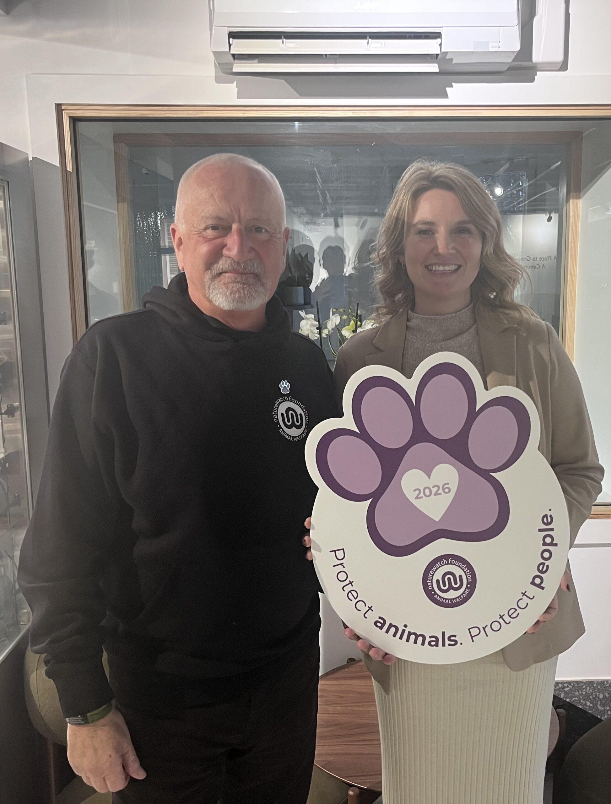 Purple Paw: Help us celebrate the special bond that we share with our pets 4 Two people stand side-by-side indoors, smiling at the camera. Mark, Naturewatch Foundation campaign manager, on the left, wears a black hoodie with the Naturewatch Foundation logo, and MP Jess Brown-Fuller, on the right, wears a beige blazer and holds a large paw-shaped sign that reads “2026 - Protect animals. Protect people.”