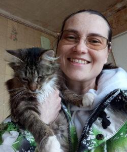 Smiling woman wearing a warm brightly patterned jacket. She is cuddling a fluffy tabby cat in an indoor setting.