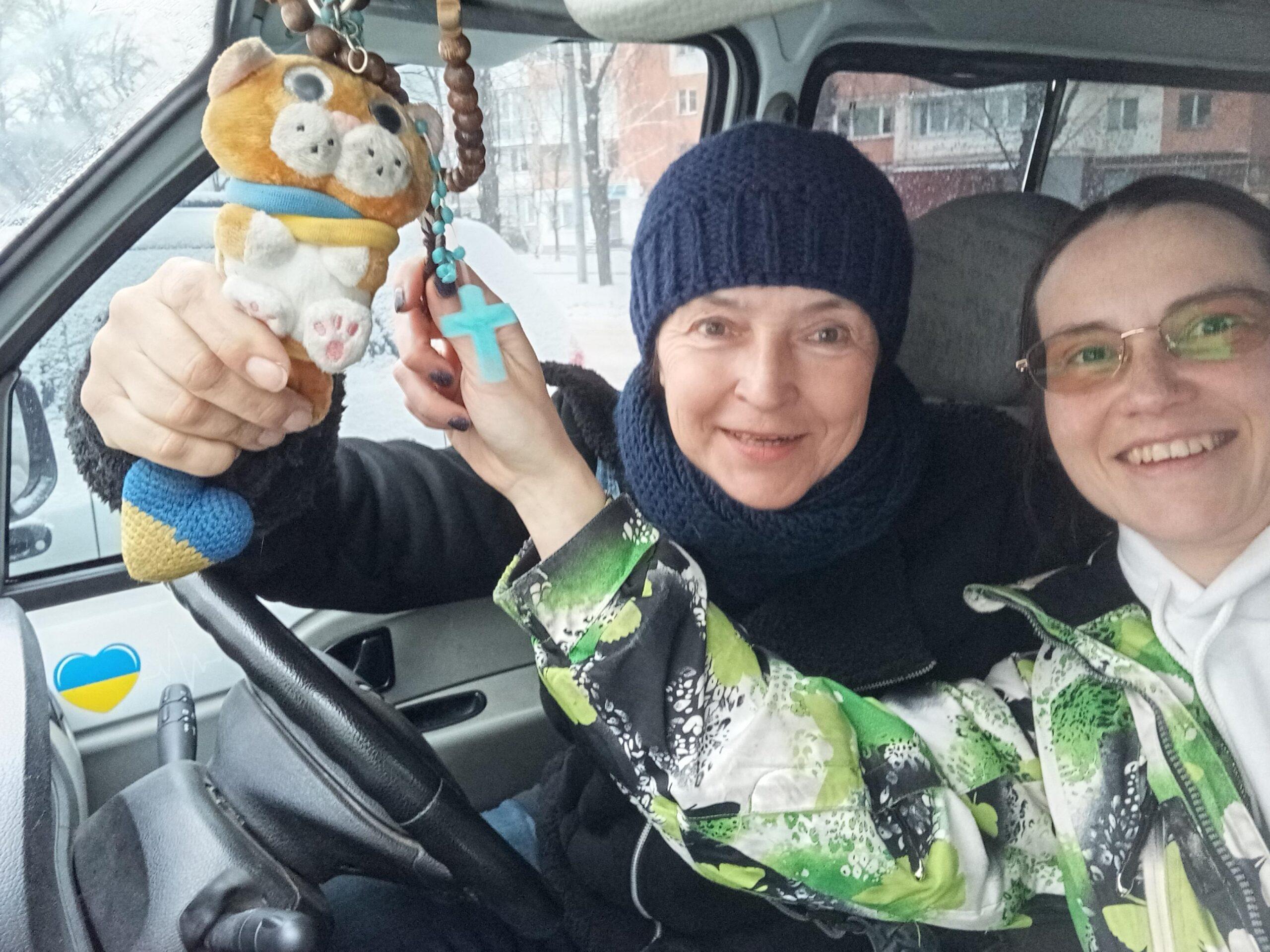 Returning to Kharkiv: Catching up with animal rescue friends in Ukraine 1 Two women sit inside a vehicle, smiling. One holds a plush toy and a cross. Both are wearing winter clothing.
