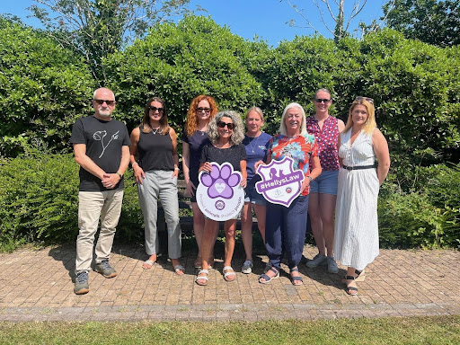 Seven people stand together outdoors in the bright sun, holding Purple Paw and Holly's Law campaign signs. They stand in front of trees and smile at the camera.
