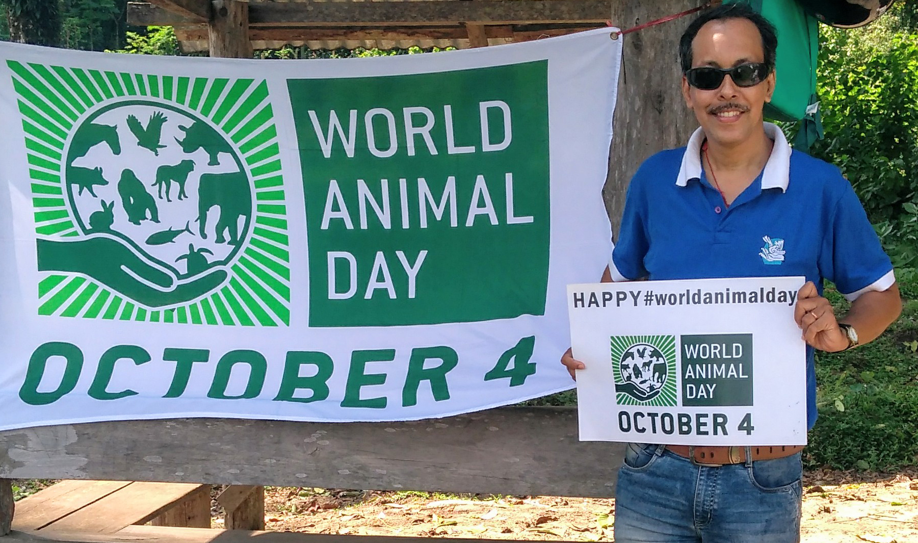 Meet the 2026 World Animal Day Grant Winners 3 A man in a blue polo shirt holds a World Animal Day sign beside a World Animal Day banner in support of People for Animals in India.