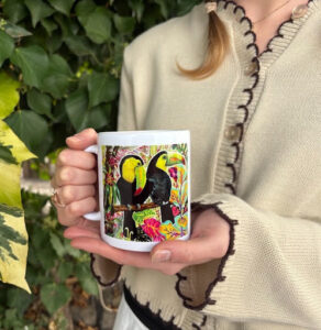A person holds the 2026 World Animal Day winning mug featuring a colourful illustration of two black and yellow toucans surrounded by tropical flowers and foliage. The photo is taken outdoors amongst a leafy backdrop