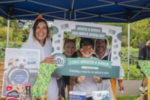A group of four people pose beneath a blue gazebo, holding a large Donate a Dinner selfie frame. On the left, a woman with long dark brown hair wears a white seagull costume. Beside her, a woman with light brown hair wears a black top and orange animal ears. A child with dark hair wears an orange-and-brown animal face mask and a white T-shirt, and a man with dark hair stands behind them in a white animal costume with an orange hood. The frame reads &ldquo;Donate a Dinner this World Animal Day&rdquo; and &ldquo;I just donated a dinner!&rdquo;