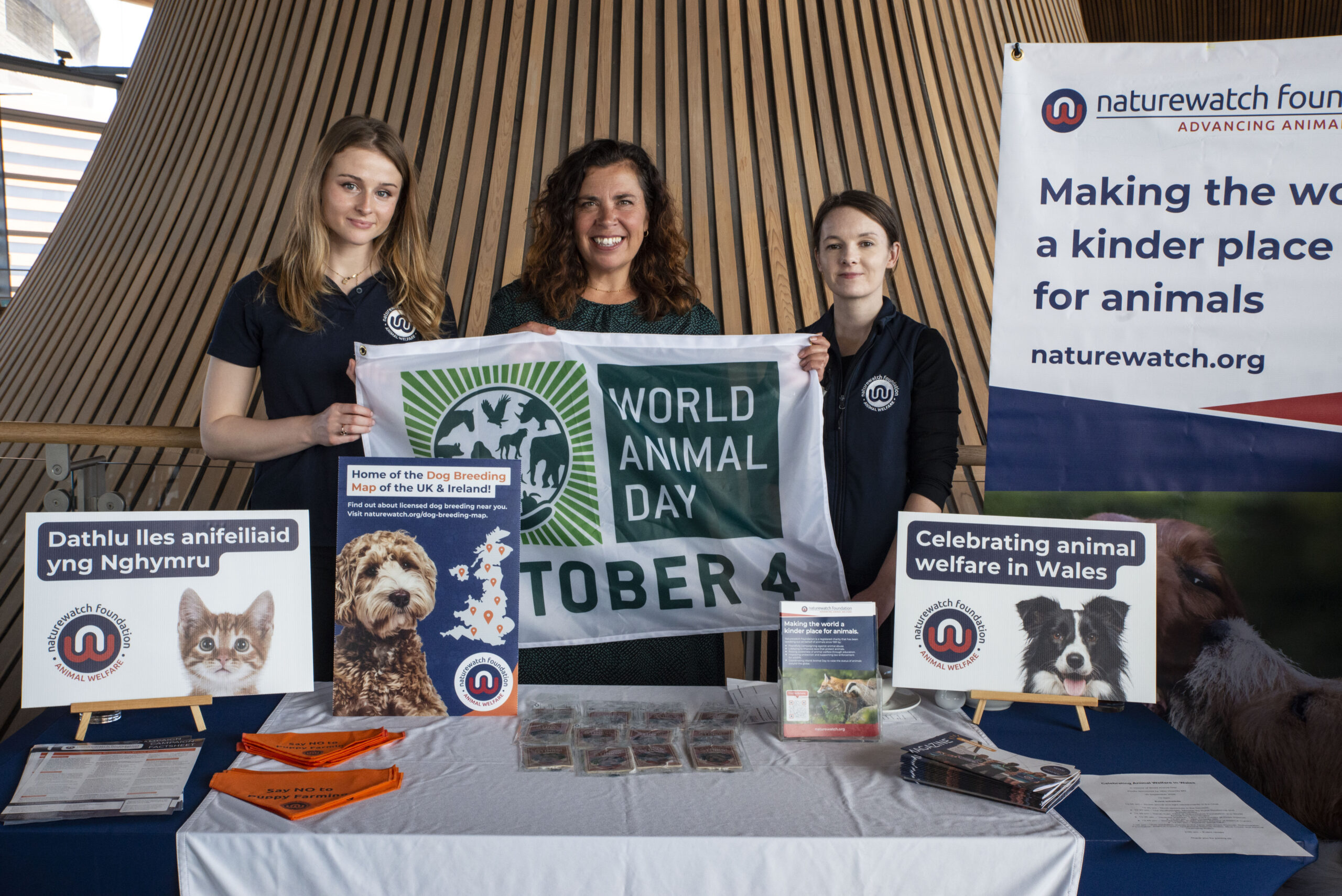 What's new 18 The Naturewatch Foundation team standing behind their table in the Senedd, holding the World Animal Day flag.