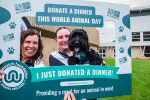 Two women and a black curly-haired dog pose with a large Donate a Dinner photo frame at Cheltenham parkrun. The frame reads “Donate a Dinner this World Animal Day” and “I just donated a dinner!”
