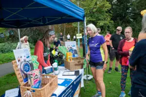 People gather at the Naturewatch Foundation Donate a Dinner stall at Cheltenham parkrun in a field surrounded by trees. The stall displays baskets of donated pet food, including boxes of cat food, along with Donate a Dinner posters, World Animal Day materials.